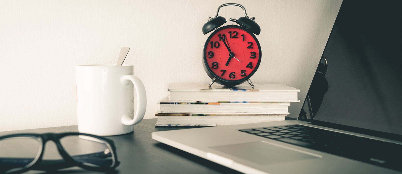 La cita sagrada con tu escritura: la disciplina es el músculo que entrena tu cerebro creativo. Un despertador rojo sobre una pila de libros junto a un ordenador portátil, gafas de lectura y una taza, simbolizando el horario fijo y el compromiso profesional del escritor.