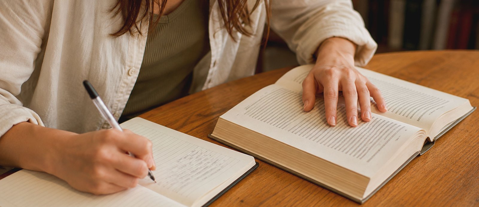 Deja de leer como un aficionado y empieza a escribir como un profesional. Primer plano de una persona tomando notas detalladas en un cuaderno junto a un libro abierto sobre una mesa de madera, representando el método práctico de la disección literaria.