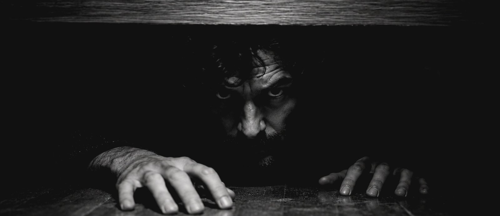 Ingeniería del suspense: cómo convertir una escena común en un momento de angustia. Imagen dramática en blanco y negro de un hombre con mirada intensa escondido en las sombras bajo un mueble, representando la tensión y el suspense literario.